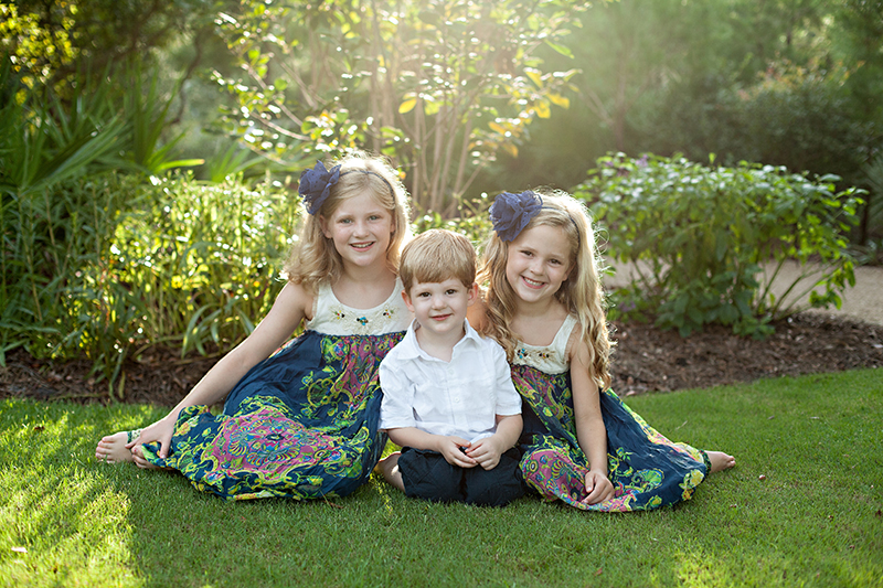 three kids posing for the camera on green lawn and the sun streaming through the trees