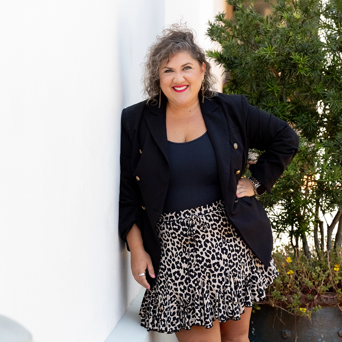Tiffany Shae - white woman with curly hair leaning against a white wall facing the camera and smiling